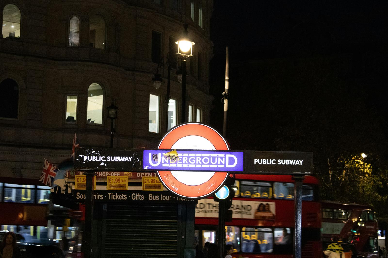 London Underground sign glowing at night with red double-decker buses passing behind